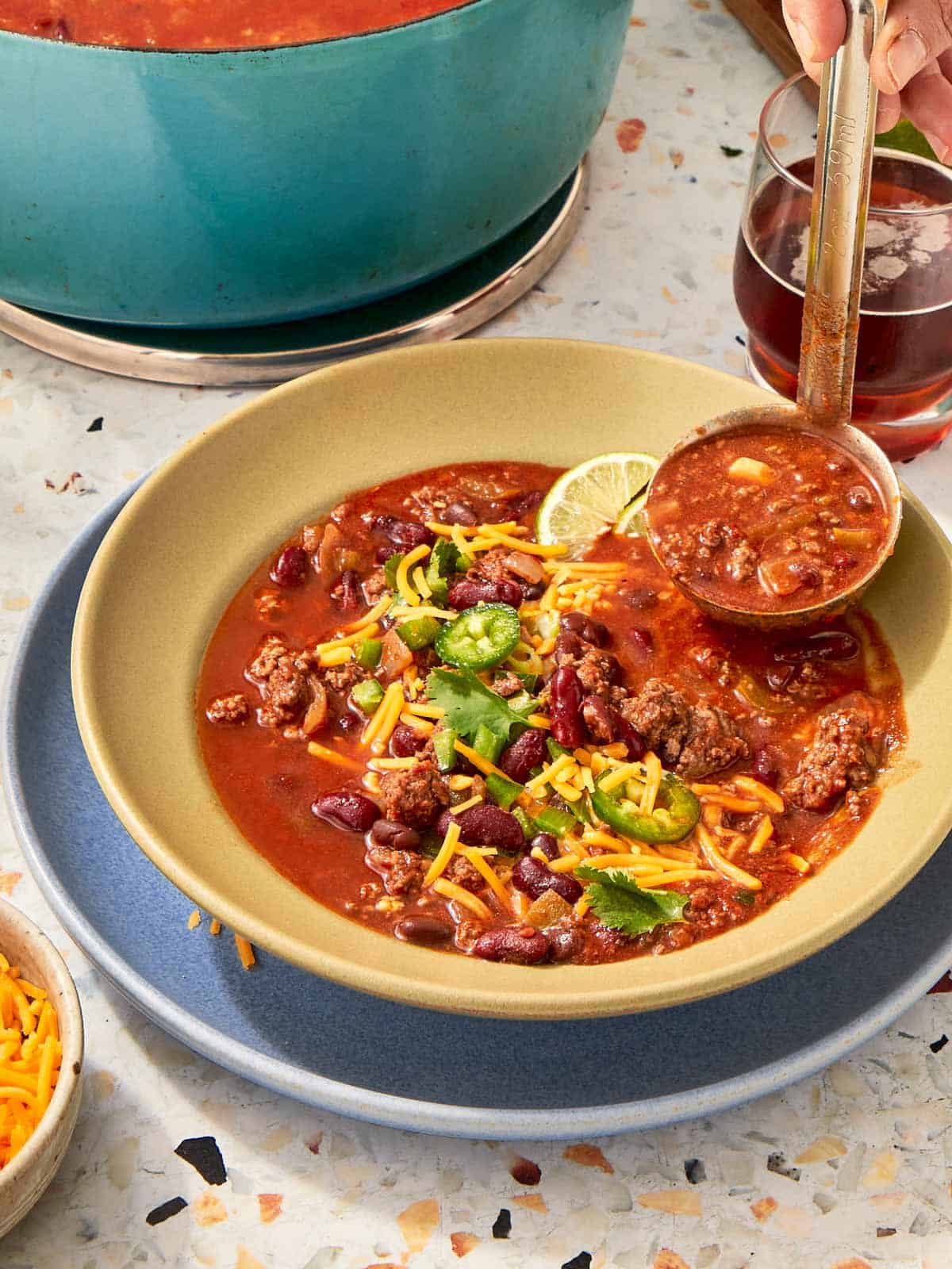 Ladling beef chili into a bowl topped with shredded cheese, cilantro, jalapeños, and lime.