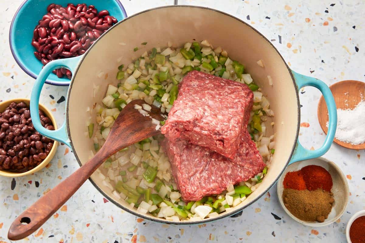 Adding raw ground beef to sautéed vegetables in a Dutch oven.