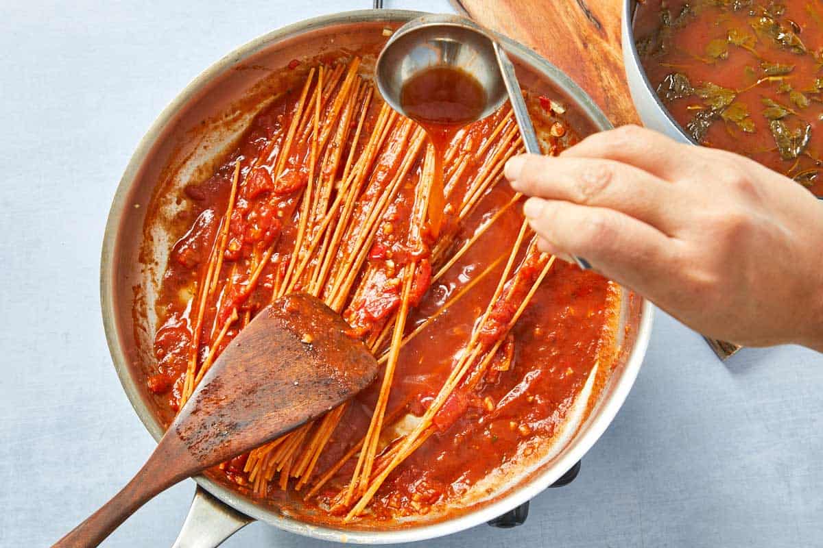 Warm broth being added spoonful by spoonful to the tomato pasta, slowly absorbed.