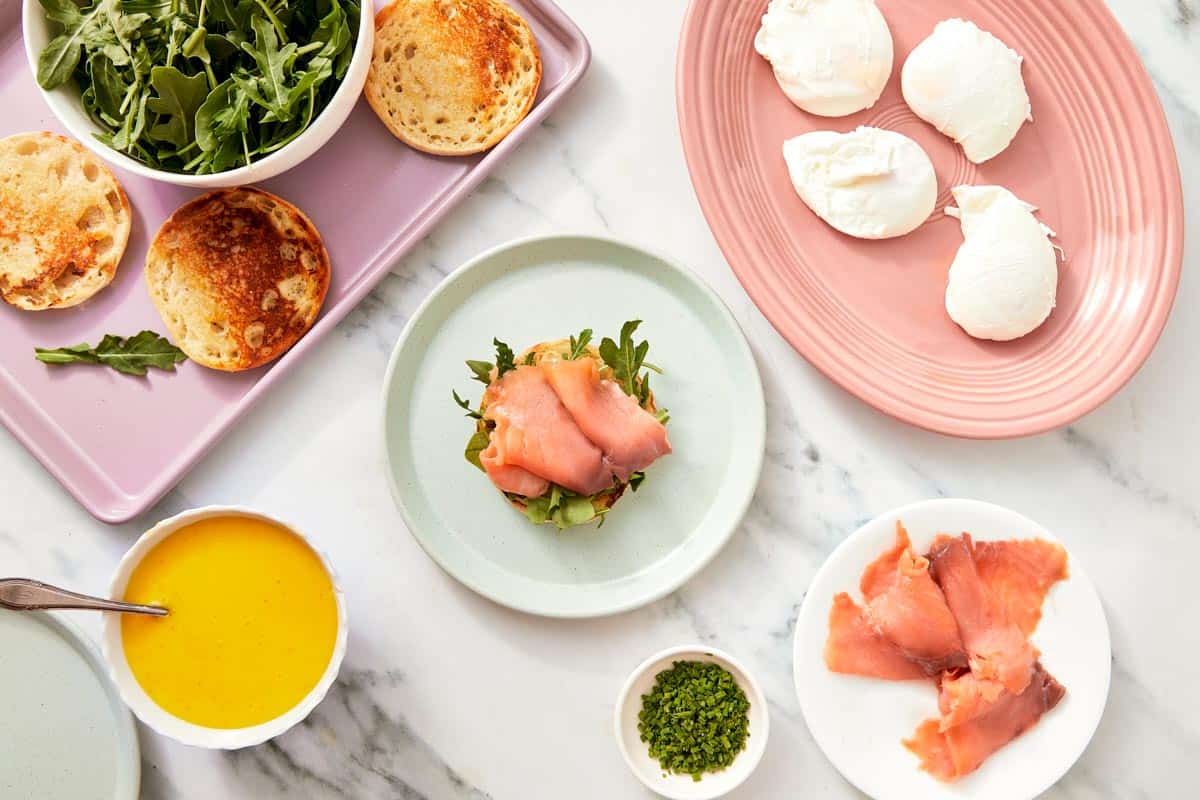 Thin slices of prosciutto arranged beside poached eggs, arugula, muffins, and hollandaise sauce on a marble surface.