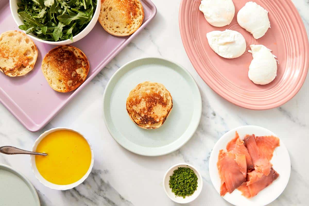 All Eggs Benedict ingredients arranged on plates: toasted muffins, poached eggs, prosciutto, arugula, hollandaise, and chives.
