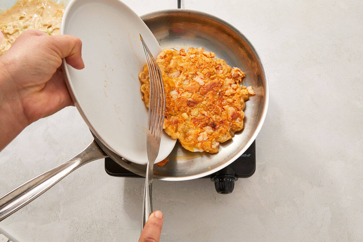 A spatula lifting the okonomiyaki as it’s being flipped in the pan with a lid held to the side.