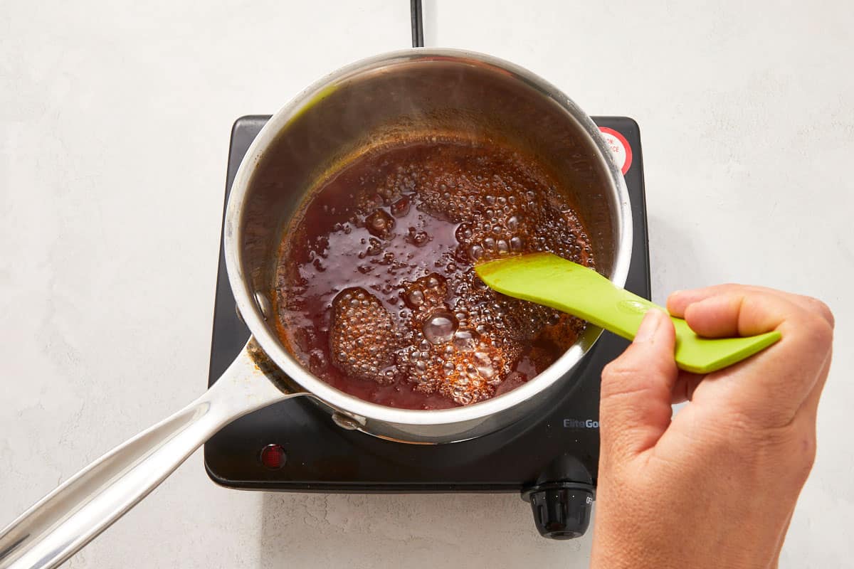 Hand stirring a dark sauce in a saucepan with a green spatula over a stovetop.