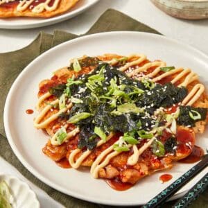 Japanese okonomiyaki pancake topped with scallions, seaweed, sesame seeds, and drizzles of mayo and sauce, served on a white plate with chopsticks.