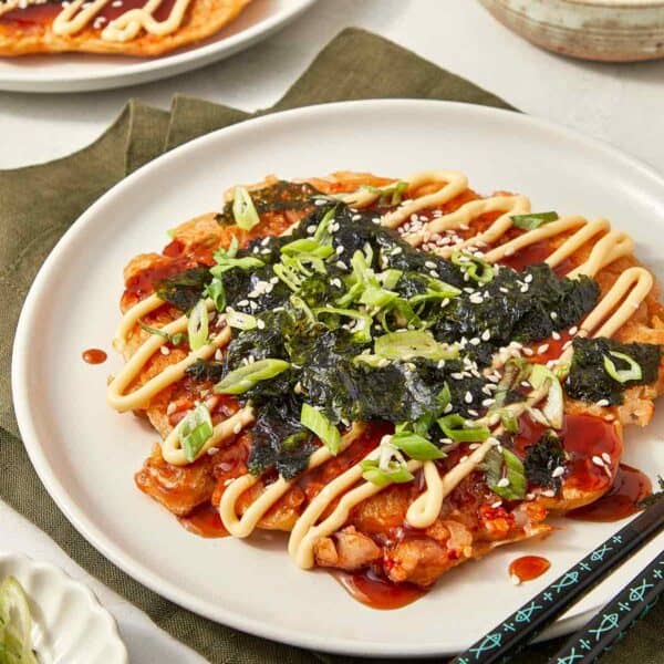 Japanese okonomiyaki pancake topped with scallions, seaweed, sesame seeds, and drizzles of mayo and sauce, served on a white plate with chopsticks.