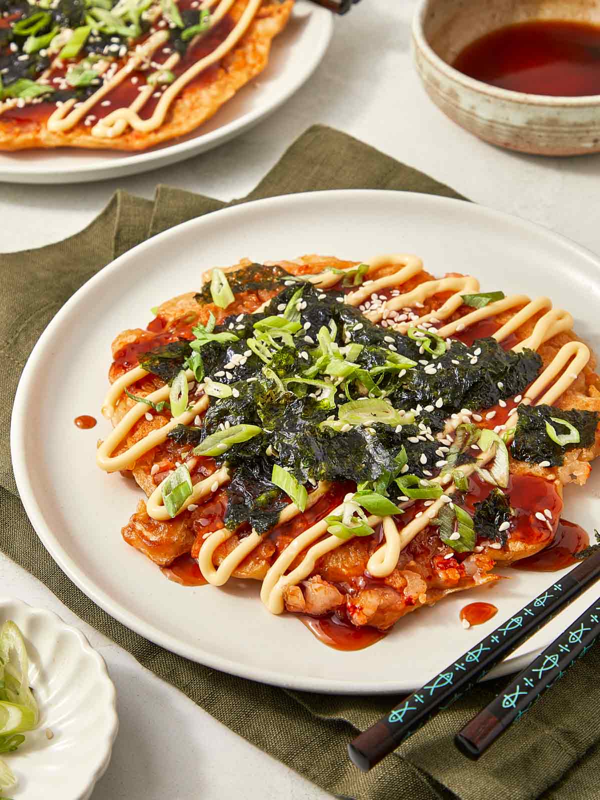Cooked okonomiyaki pancake topped with drizzles of mayo and sauce, garnished with chopped scallions, sesame seeds, and shredded seaweed on a white plate with chopsticks.