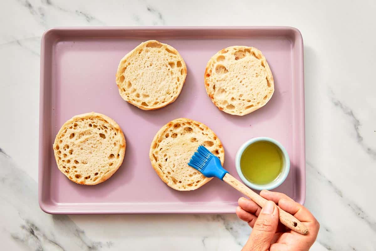 A hand brushing English muffin halves with olive oil on a pink tray.