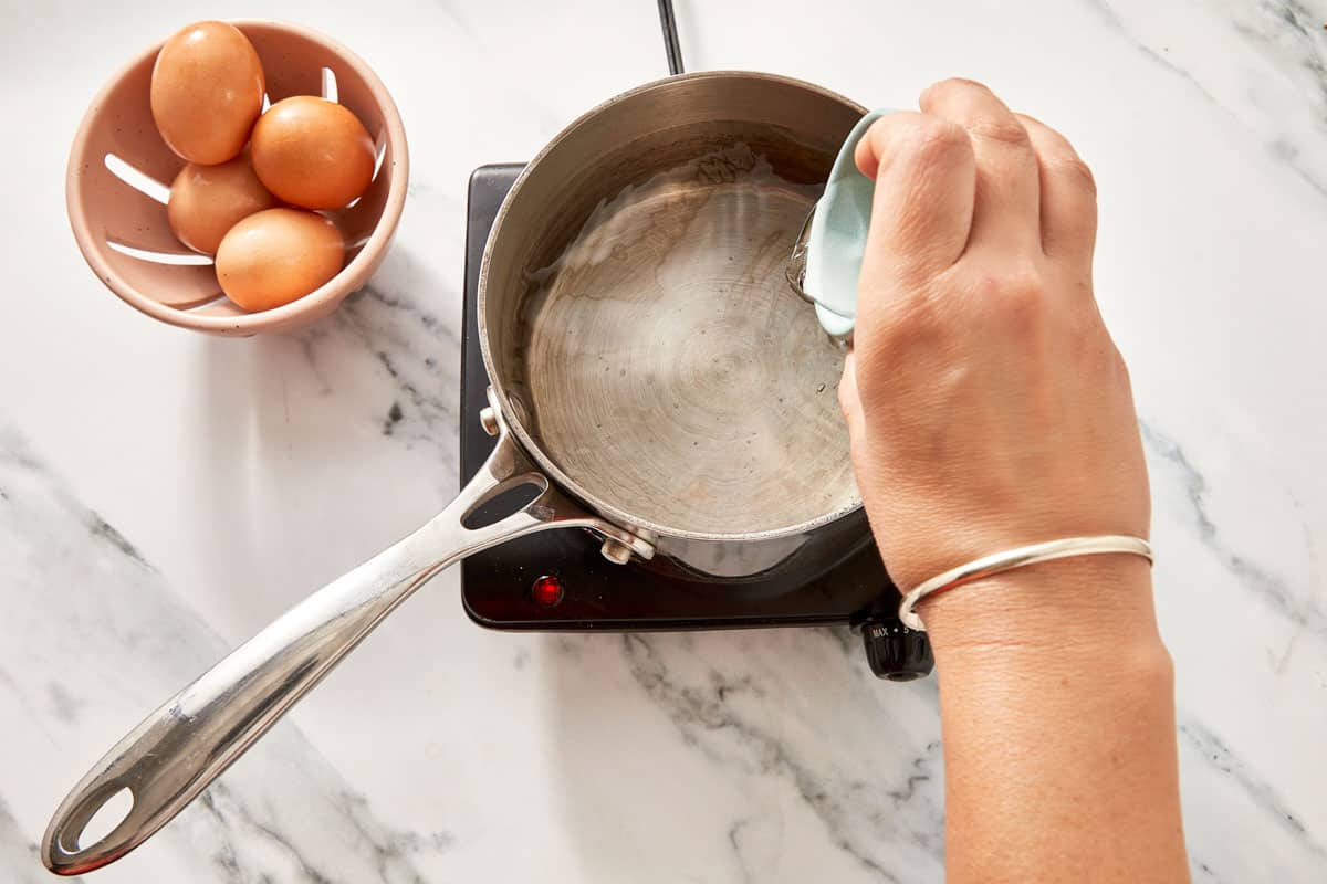 A hand holding a ramekin over a saucepan with gently simmering water and a spoon creating a whirlpool.