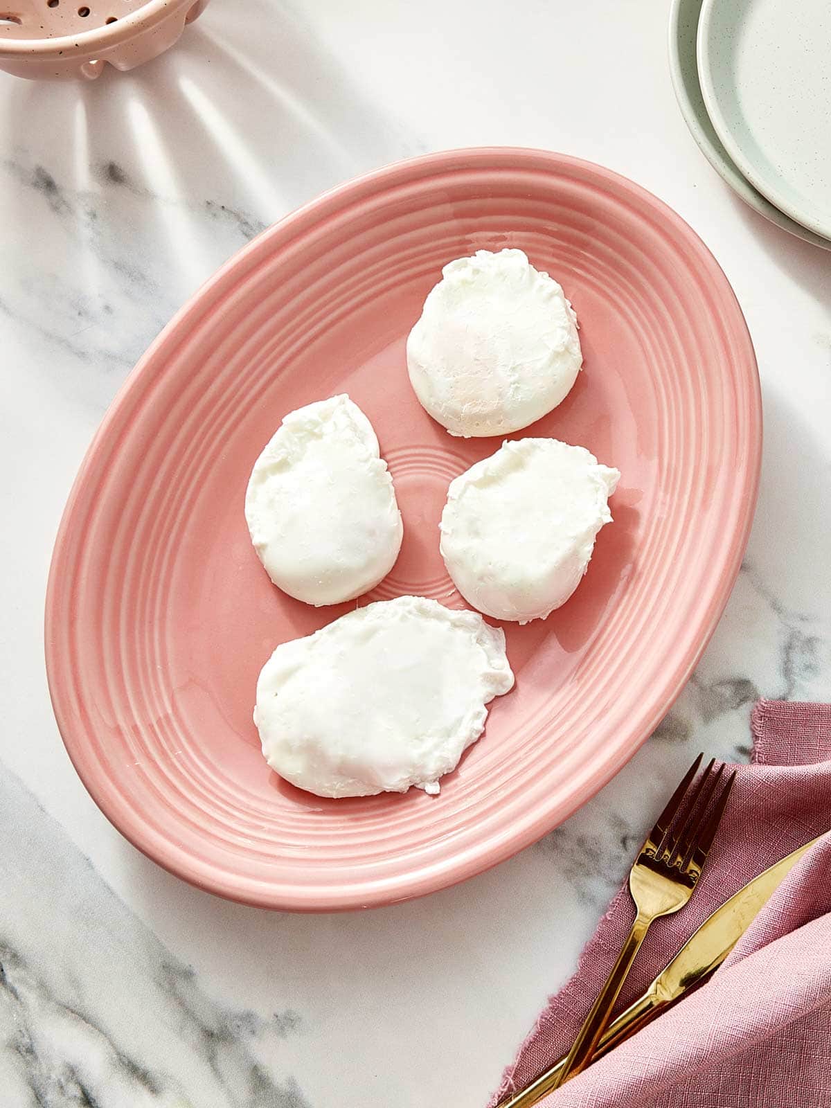 Four perfectly poached eggs resting on a blush-pink plate over a marble background.