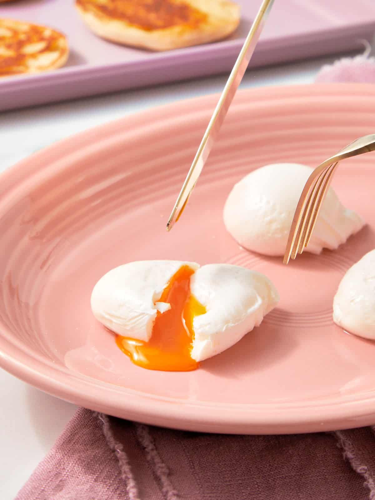 Three poached eggs on a plate, with a fork and knife cutting into one — the golden yolk spilling out softly.