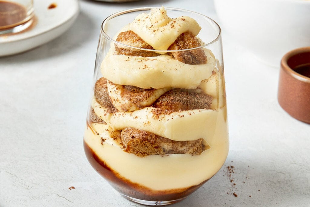 Single serving of tiramisu in a glass, layered with creamy mascarpone and espresso-soaked ladyfingers, topped with cocoa powder.