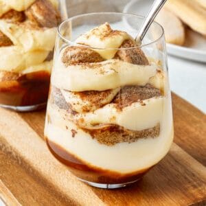 Two tiramisu cups served in clear glasses on a wooden board, layered with mascarpone cream, ladyfingers, and a dusting of cocoa.