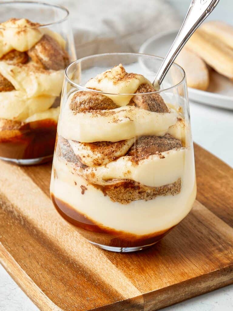 Two tiramisu cups served in clear glasses on a wooden board, layered with mascarpone cream, ladyfingers, and a dusting of cocoa.