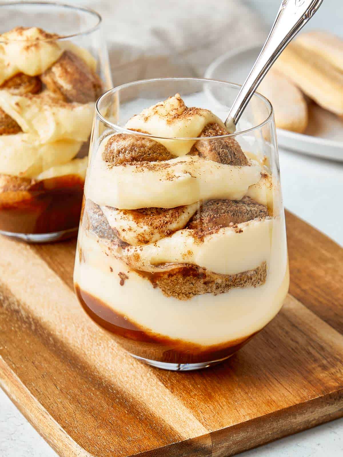 Two tiramisu cups served in clear glasses on a wooden board, layered with mascarpone cream, ladyfingers, and a dusting of cocoa