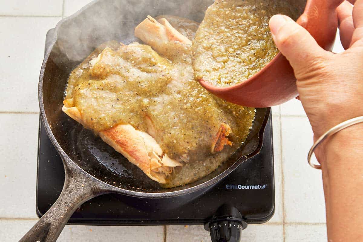 A cook adds green salsa to enchiladas in a skillet, ready to cover and simmer for seven minutes.