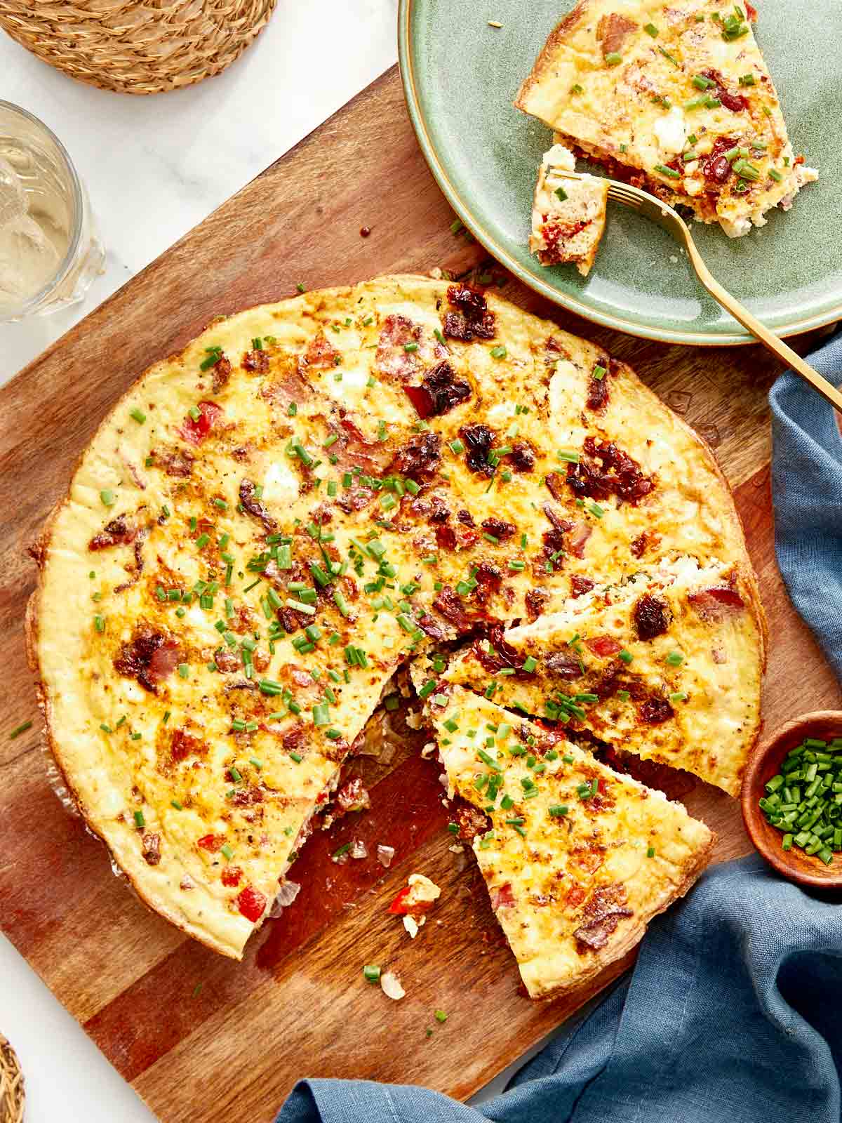 Whole frittata on a wooden board, golden and puffed, topped with crispy bacon and chives, surrounded by serving plates and flatware.