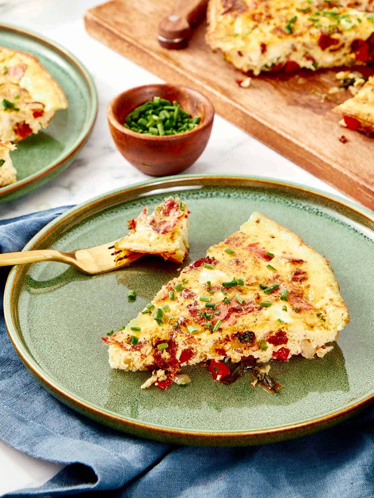A slice of frittata served on a green ceramic plate, garnished with fresh chives, with a forkful ready to eat.