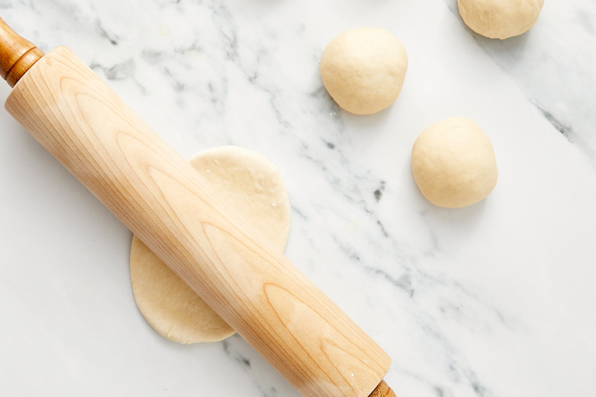 Small bao dough balls being rolled into ovals with a wooden rolling pin.