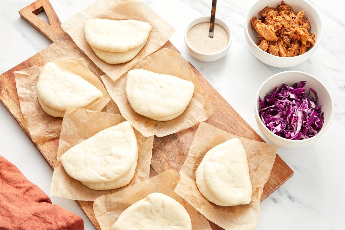 Steamed bao buns ready to be filled, next to bowls of pulled pork, shredded cabbage, and black garlic mayo.