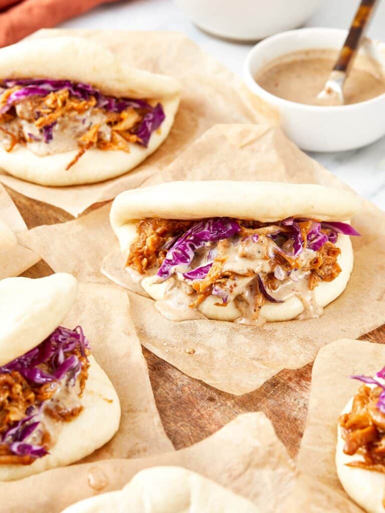 Steamed bao buns filled with pulled pork, red cabbage, and drizzled black garlic mayo on parchment paper.