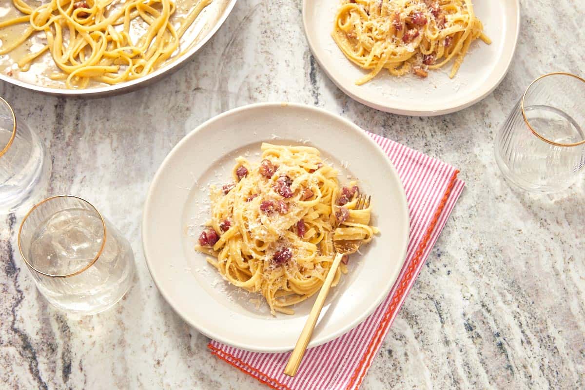 A plate of finished pasta carbonara on a red-striped napkin, topped with crispy pancetta and extra cheese.