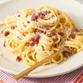A plate of finished pasta carbonara on a red-striped napkin, topped with crispy pancetta and extra cheese. - 11