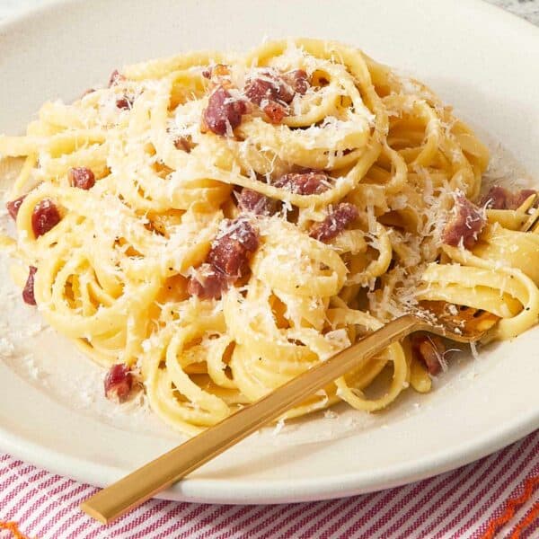 A plate of finished pasta carbonara on a red-striped napkin, topped with crispy pancetta and extra cheese.