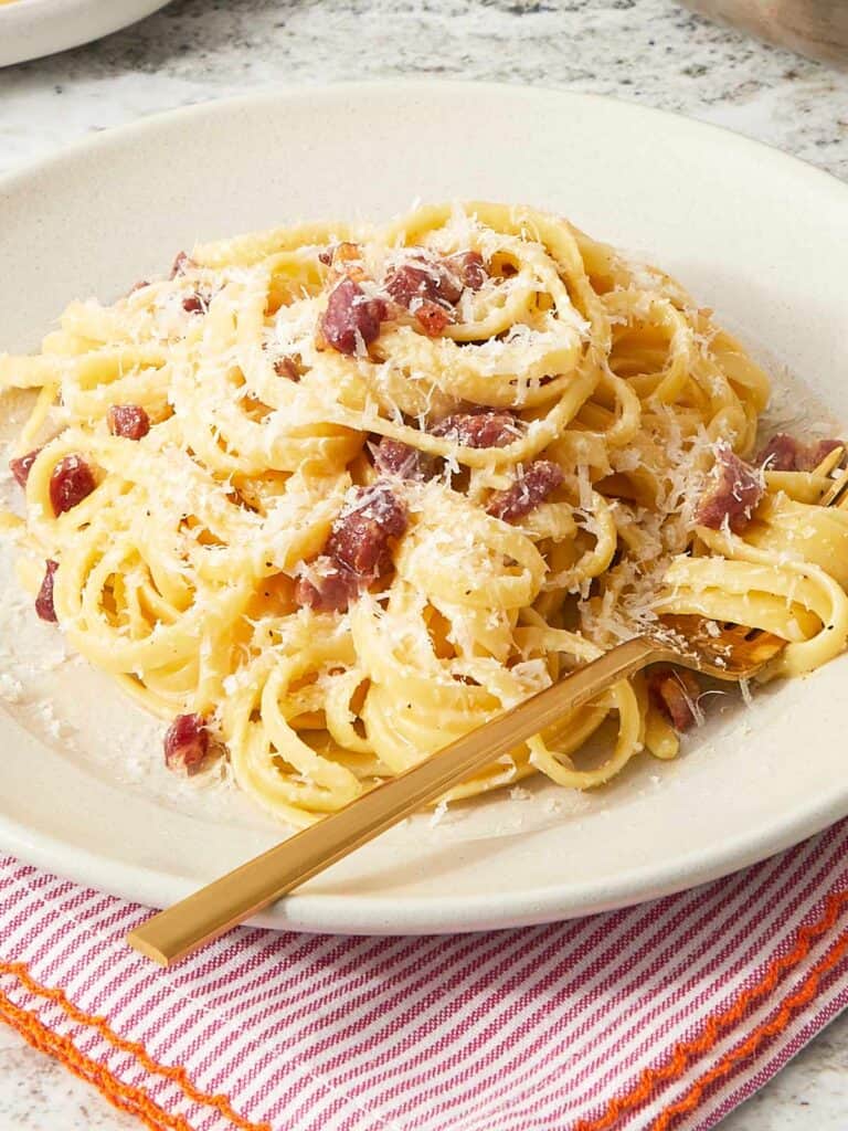 A plate of finished pasta carbonara on a red-striped napkin, topped with crispy pancetta and extra cheese.