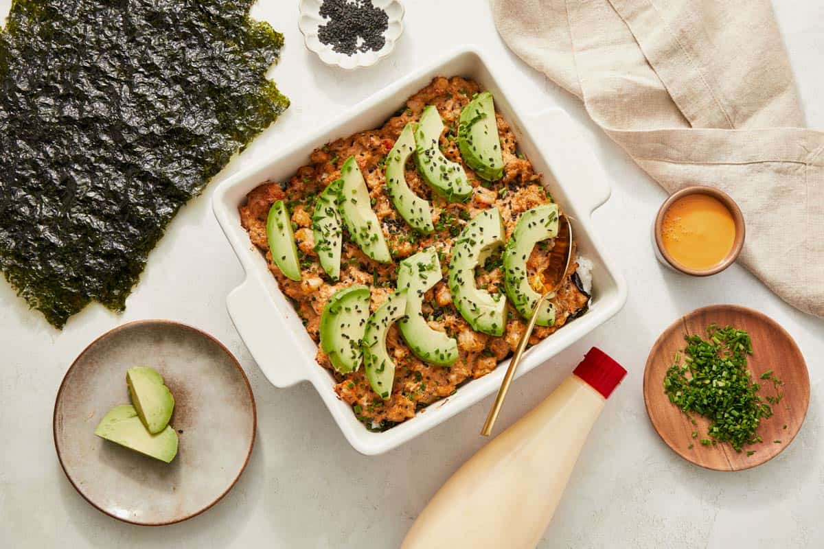 A cooked sushi bake casserole featuring avocado, green beans, and various other ingredients, served in a baking dish.

