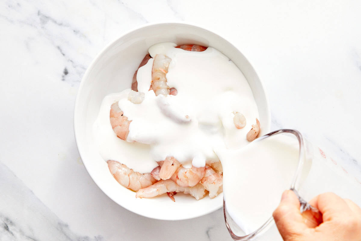 Raw shrimp in a white bowl with buttermilk being poured over them for marinating.