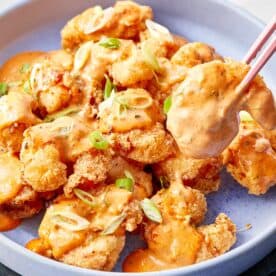Shrimp coated in creamy bang bang sauce, garnished with sliced green onions, served in a blue bowl with chopsticks lifting one piece.