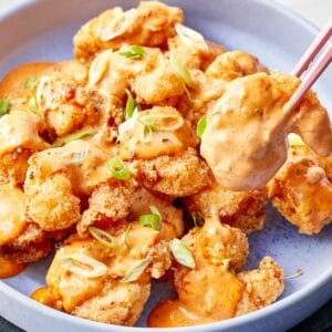 Shrimp coated in creamy bang bang sauce, garnished with sliced green onions, served in a blue bowl with chopsticks lifting one piece.