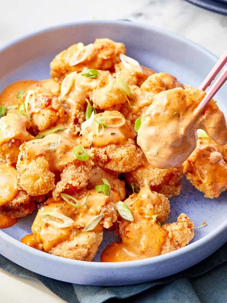 Shrimp coated in creamy bang bang sauce, garnished with sliced green onions, served in a blue bowl with chopsticks lifting one piece.