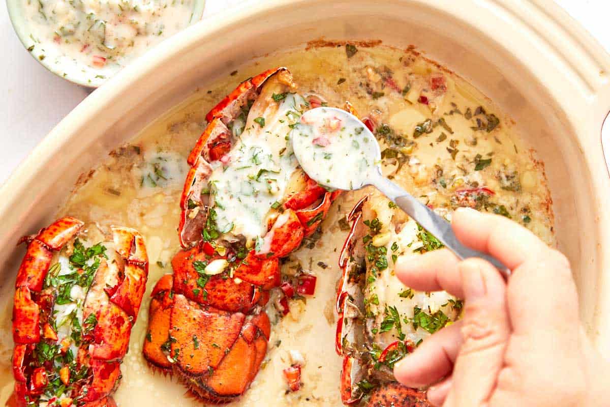 Close-up of chimichurri butter being spooned onto broiled lobster tails in a baking dish.