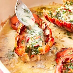 Close-up of a lobster tail being drizzled with creamy chimichurri butter, highlighting the vibrant herbs and rich sauce pooling in the baking dish.