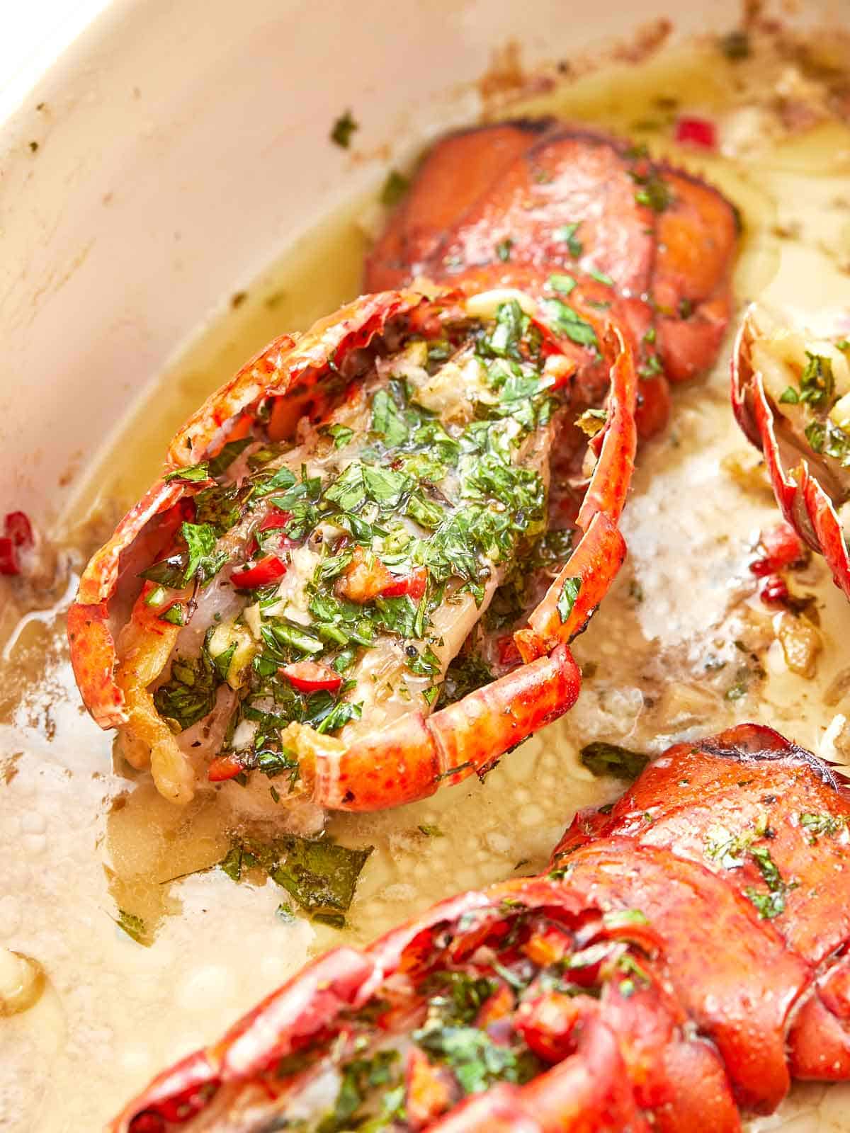 Close-up of a broiled lobster tail topped with chimichurri butter, herbs, and red chili, sitting in a buttery pan sauce.