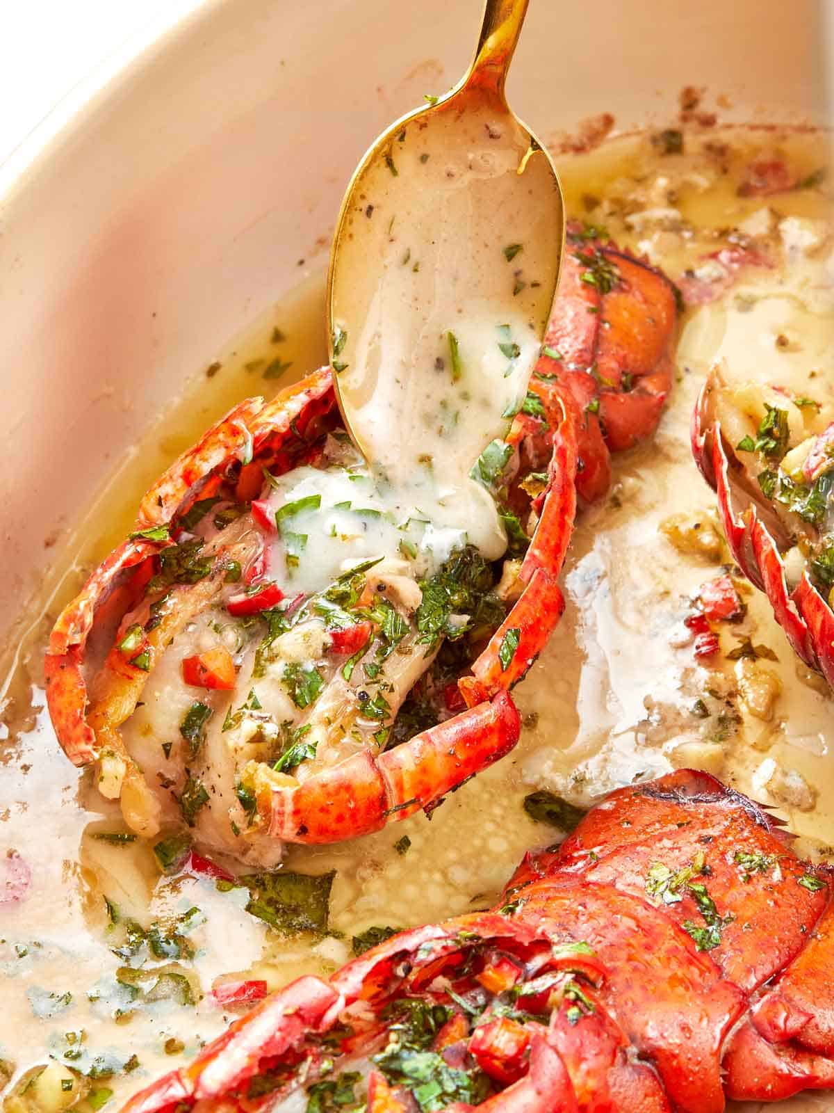 Close-up of a broiled lobster tail topped with chimichurri butter, herbs, and red chili, sitting in a buttery pan sauce.
