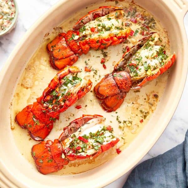 Four broiled lobster tails in a baking dish, topped with chimichurri butter and garnished with fresh herbs and red chili.