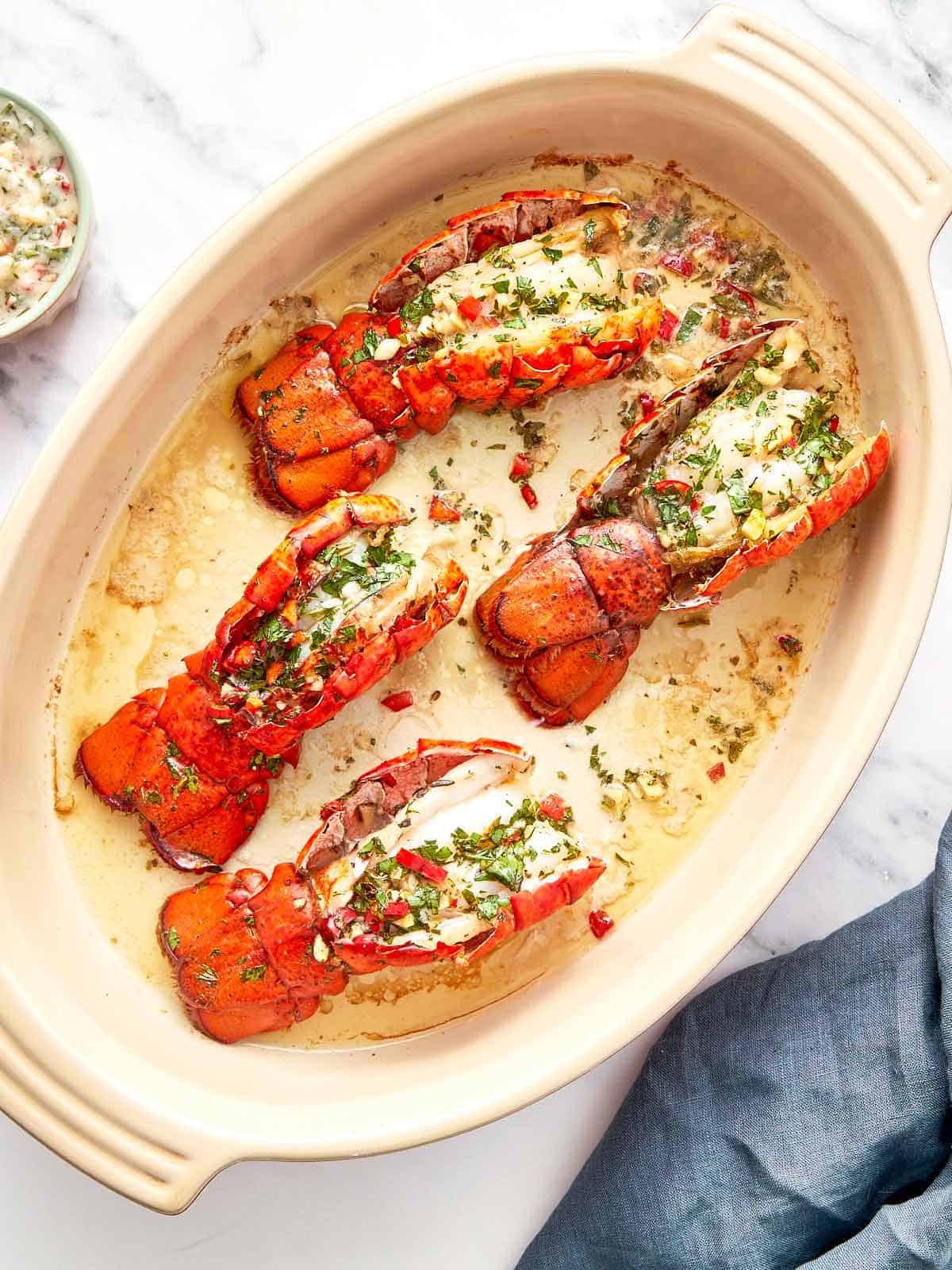 Four broiled lobster tails in a baking dish, topped with chimichurri butter and garnished with fresh herbs and red chili.