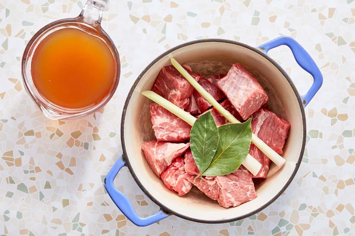 Beef chunks with lemongrass and bay leaf in a pot next to beef broth.