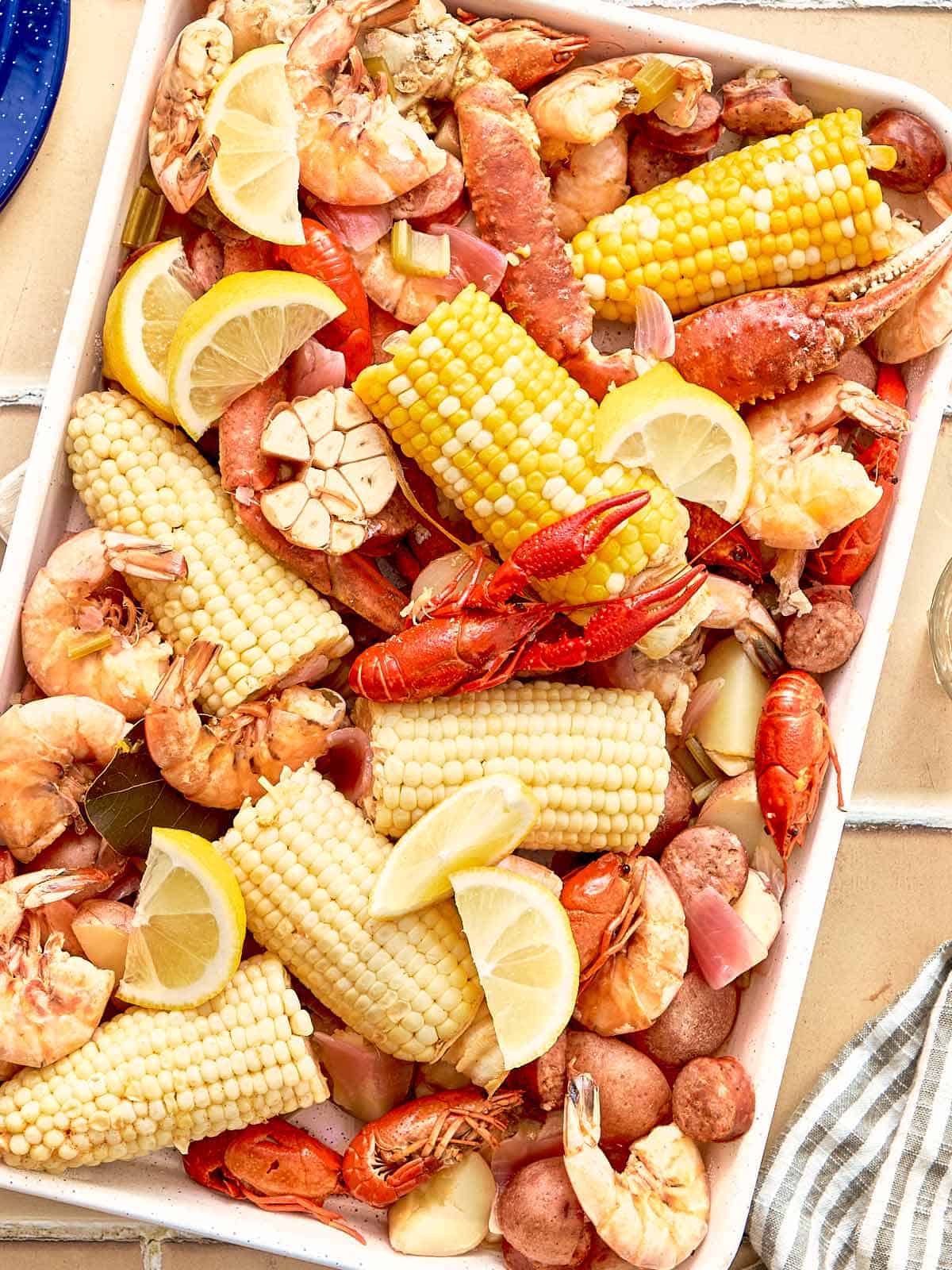 Top-down view of a cooked seafood boil tray with corn, shrimp, crab legs, crawfish, potatoes, sausage, garlic, and lemon wedges.