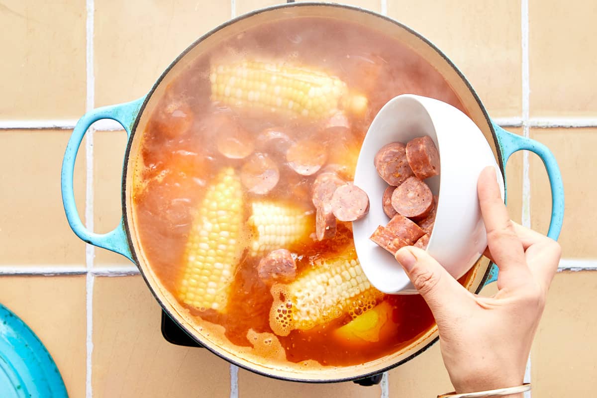 Corn boiling in the broth while a hand adds sliced sausage into the pot.
