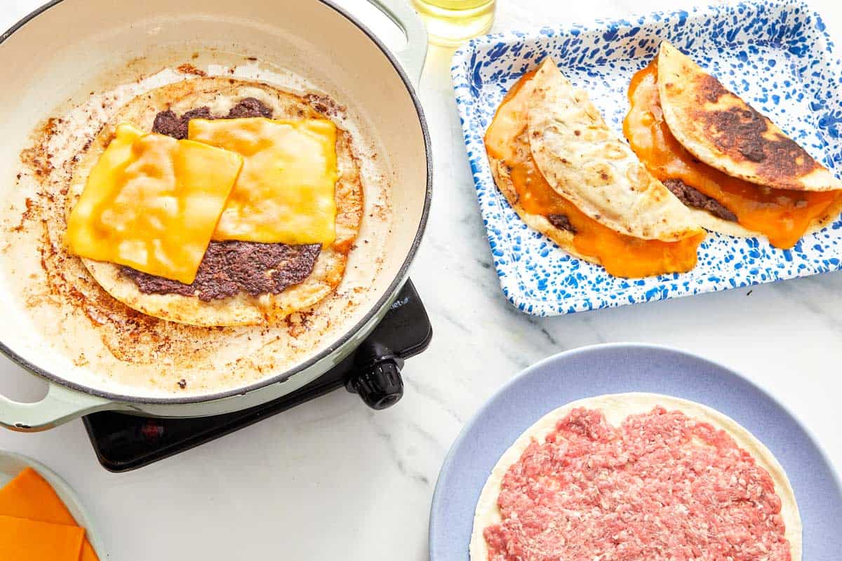 Cooking beef-topped tortillas with melted cheddar cheese in a skillet, with finished cheesy tacos on the side."