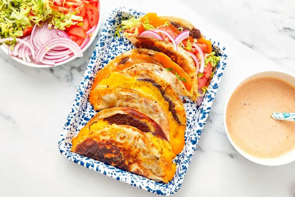 Tray of cheesy smash burger tacos with lettuce, tomato, and red onion, served with burger sauce on the side.