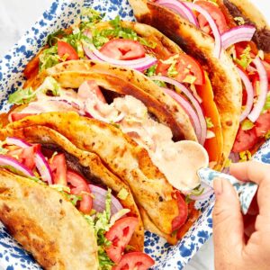 Close-up of smash burger tacos in a blue speckled tray, filled with melted cheese, lettuce, tomato, and red onion, being topped with creamy sauce.