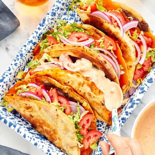 Smash burger tacos in a blue speckled tray, filled with beef, melted cheese, shredded lettuce, sliced tomatoes, red onions, and topped with creamy sauce.