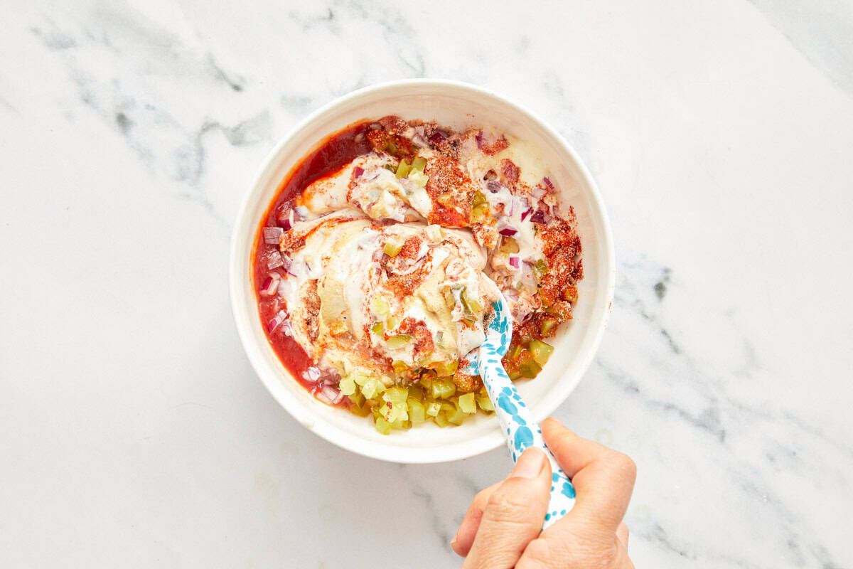 Bowl with mayonnaise, ketchup, chopped pickles, diced red onion, and spices being mixed with a spoon.
