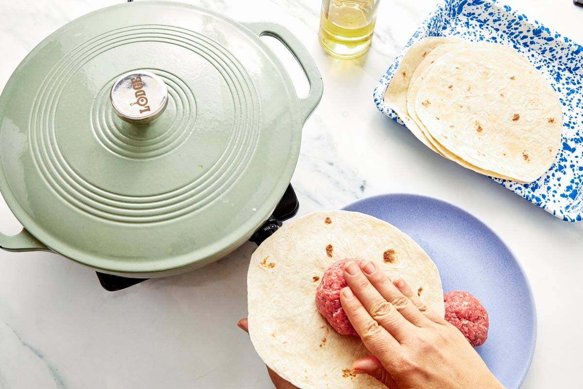 Pressing a raw beef ball onto a flour tortilla before cooking in a skillet.