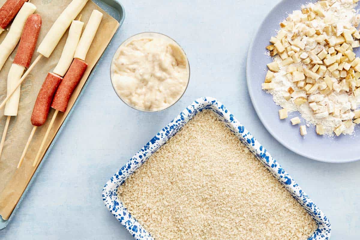 Prep station with batter, panko breadcrumbs, and diced fries ready for coating corn dogs.