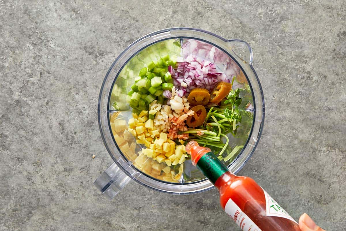 Blender filled with fresh ginger, garlic, celery, onion, coriander stems, jalapeño, and hot sauce.